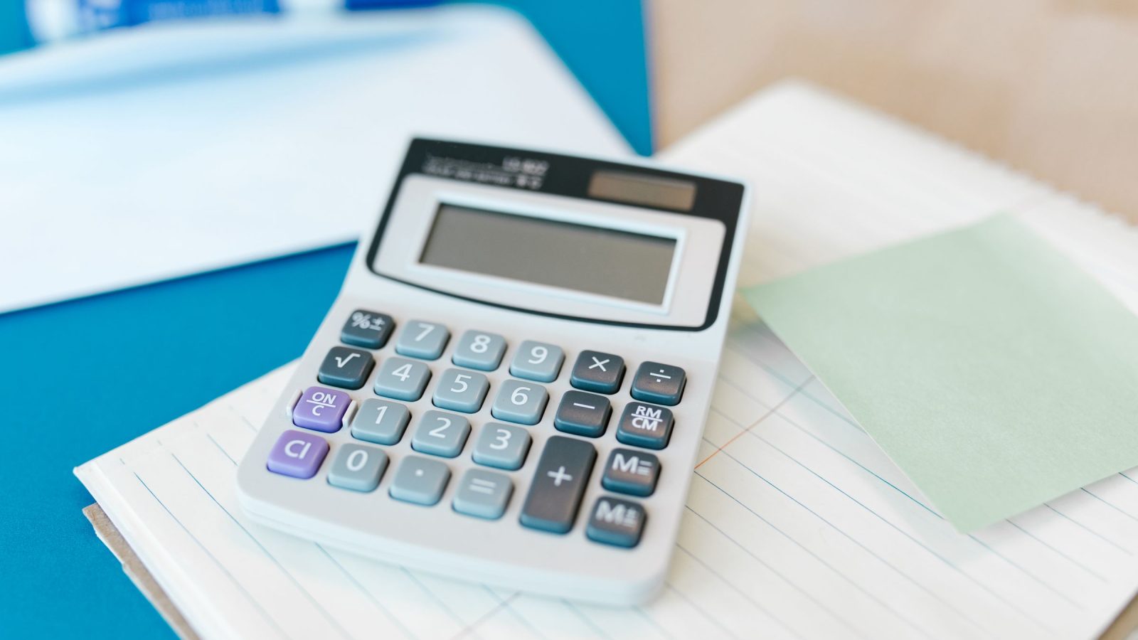 Calculatrice posée sur un bloc-notes et des feuilles, avec un pense-bête vert sur fond bleu