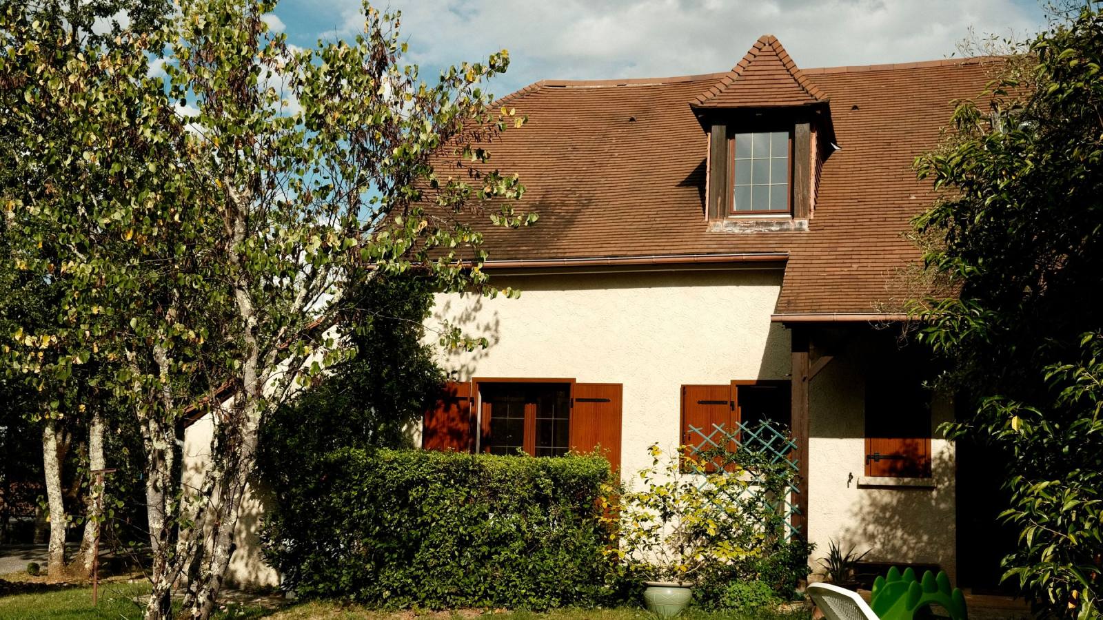 Maison au toit de tuiles brunes, façade en crépi crème, volets bois et lucarne, entourée de verdure