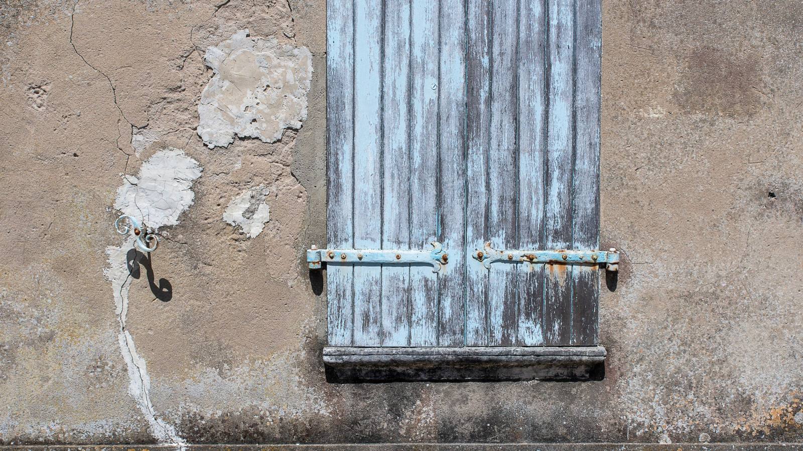Volet en bois bleu pâle à la peinture écaillée, sur un mur d'enduit usé et fissuré