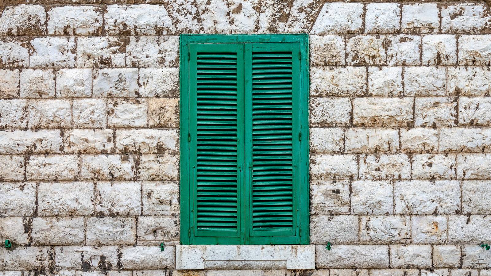 Fenêtre aux volets persiennes verts sur un mur en pierres claires appareillées