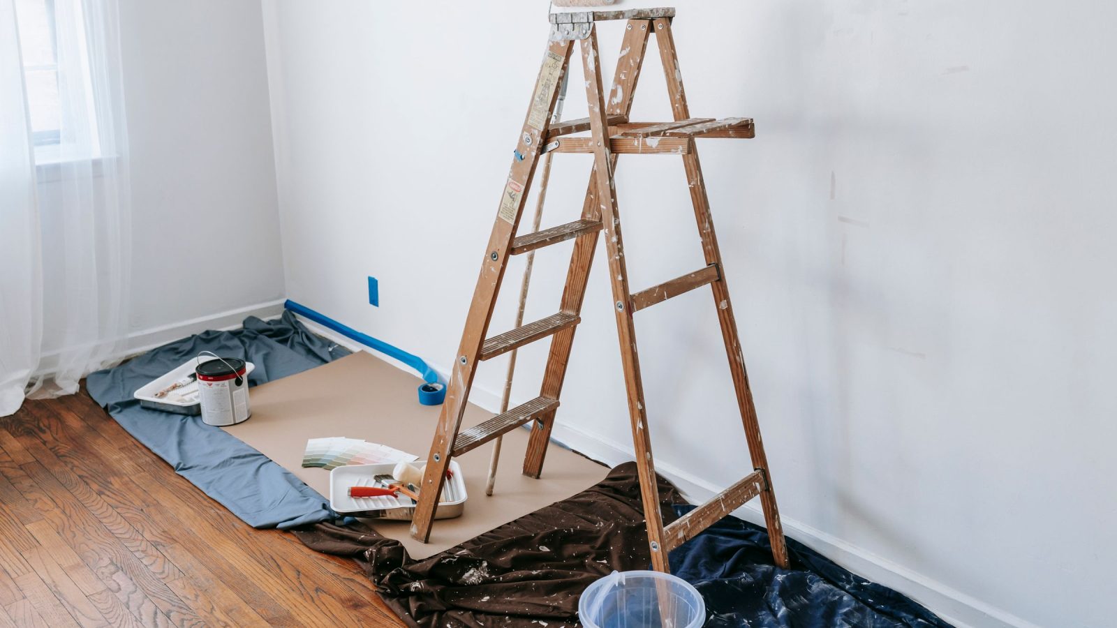 Pièce en cours de peinture : escabeau en bois, bâche au sol, pots et pinceaux contre un mur blanc