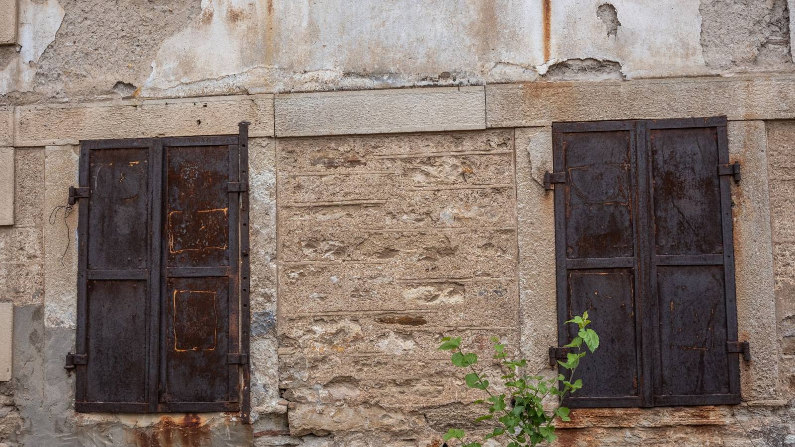 Deux volets métalliques sombres piqués de rouille sur une façade de pierre et d'enduit patiné